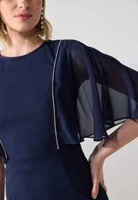 Robe bleu marine avec des manches en dentelle flottantes et une rangée de décorations argentées sur le devant pour plus de détails et de texture.