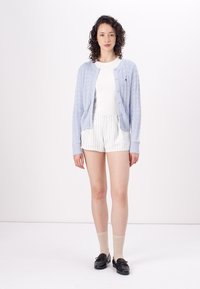 Light blue knitted cardigan with cable pattern, button closure, worn over a white top and striped shorts. Black shoes and light beige socks.