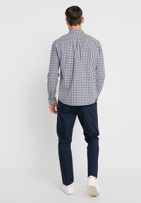 Uomo in piedi di spalle, che indossa una camicia a scacchi bianca e nera, pantaloni blu scuro e sneaker bianche, su uno sfondo semplice.