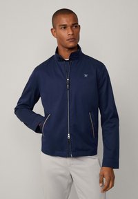 Chaqueta azul marino con cremallera frontal, cuello alto, dos bolsillos laterales con detalles en marrón claro y un pequeño logo en el pecho. Textura de tela suave.