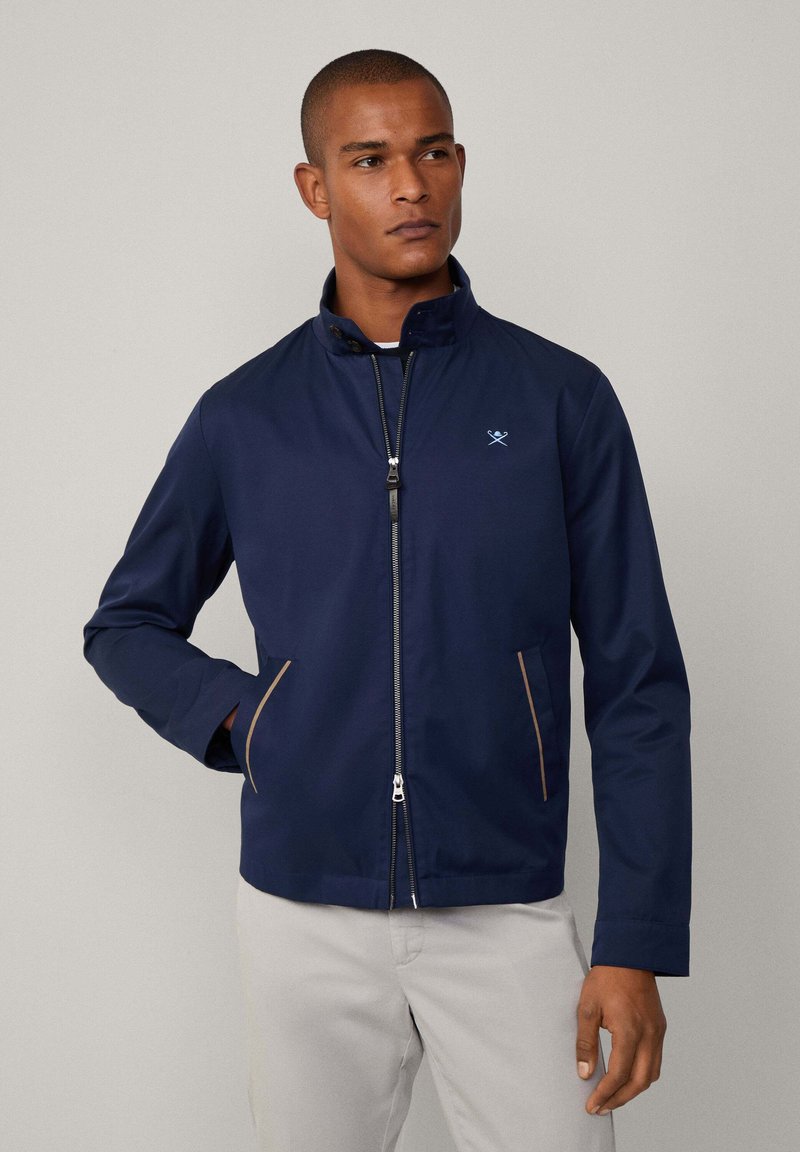 Chaqueta azul marino con cremallera frontal, cuello alto, dos bolsillos laterales con detalles en marrón claro y un pequeño logo en el pecho. Textura de tela suave.
