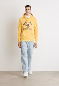 Sweat-shirt à capuche jaune avec "GANT 1949 SPORTSWEAR" imprimé, assorti à un jean droit bleu clair et des baskets blanches.