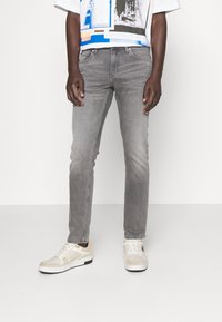 Man som bär ljusgrå slim-fit jeans, vita sneakers med svarta sulor, och en del av en vit grafisk t-shirt mot en enkel vit bakgrund.