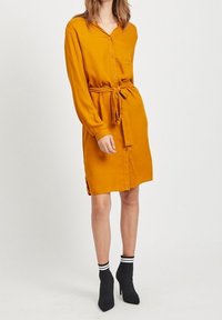 Robe chemise jaune moutarde à manches longues, devant boutonné, taille ceinturée et fentes latérales. Associée à des bottines à talons noires avec des rayures blanches.