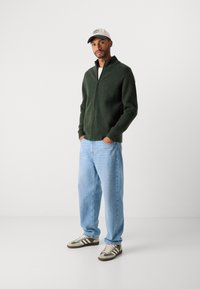 Mann med hvit og grønn caps, mørkegrønn zip-up hettegenser, lyseblå jeans og grønn og hvit sneakers, står med hendene i lommene.