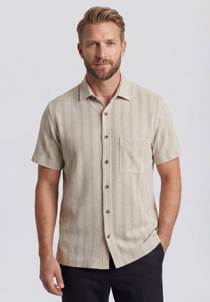 Homme blond avec une barbe courte porte une chemise beige à manches courtes avec des rayures verticales subtiles et une poche avant, debout avec une main dans la poche.