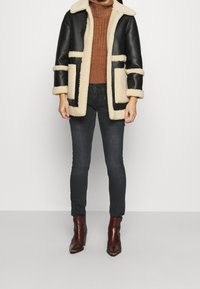 Musta nahkainen karvahousutakki, jossa on kermahtavat reunukset, kaksirivinen malli ja etutaskut, päällä ruskea ribbineulottu villapaita ja tummat farkut.