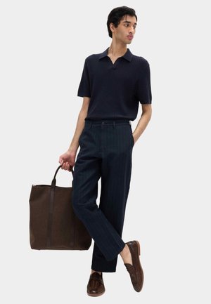 Joven con polo de manga corta acanalado en azul marino y pantalones de rayas, llevando un gran bolso tote marrón, calzando zapatos marrones de cuero trenzado.
