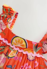 Klänning i lysande orange med volangband, rosa vågig kant och ett mönster med citrusfrukter, inklusive citroner och andra färgglada frukter.