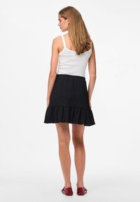 Donna con top bianco senza maniche, minigonna nera con balze e scarpe rosse basse, in piedi di spalle su sfondo bianco.