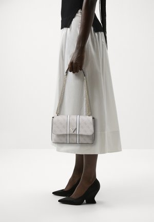 Vrouw in witte rok en zwarte top houdt een witte Guess handtas met zilveren kettingriem vast, draagt zwarte pumps met puntige neus.