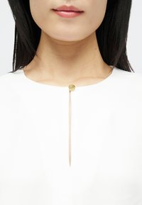 Žena v bílém topu zapnutém na krku jediným kulatým zlatým knoflíkem, ukazující část její dolní tváře a krku.