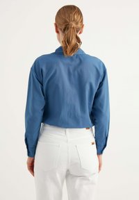 Camicia azzurra a maniche lunghe con colletto, texture liscia e polsini abbottonati, indossata con pantaloni bianchi a vita alta con tasche posteriori.