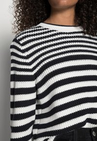 Sweter dzianinowy w czarno-białe paski, z okrągłym dekoltem, teksturowanym materiałem i luźnym fasonem, z widocznymi poziomymi paskami.