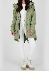 Olivgrön parka med dragkedja och tryckknappsstängning, justerbar huva och två framfickor. Kombinerad med vita åtsittande byxor och kraftiga vita stövlar.