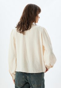 Blusa color crema con finitura testurizzata, maniche a sbuffo e taglio dritto. Indossata con jeans di denim a vita alta.