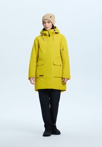 Parka gialla impermeabile con cappuccio, zip frontale, due grandi tasche e coulisse regolabili. Indossata con pantaloni neri e stivali.