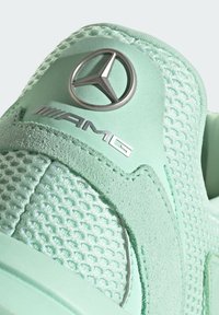 Zelená sportovní bota zblízka, která se vyznačuje texturovaným síťovaným svrškem a hladkými akcenty, se stříbrným logem AMG a kruhovým emblémem Mercedes.