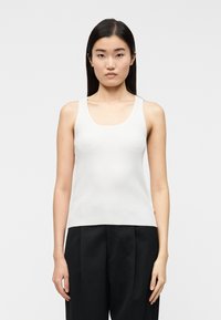 Γυναίκα που στέκεται με το σώμα προς τα εμπρός, φοράει άσπρο ριμπ τοπ χωρίς μανίκια και μαύρο παντελόνι, μπροστά σε ένα απλό φωτεινό φόντο.