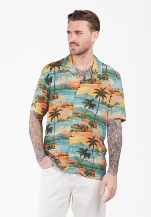 Hombre con tatuajes que lleva una camisa de manga corta con estampado tropical colorido y pantalones blancos, mirando de lado con las manos parcialmente en los bolsillos.