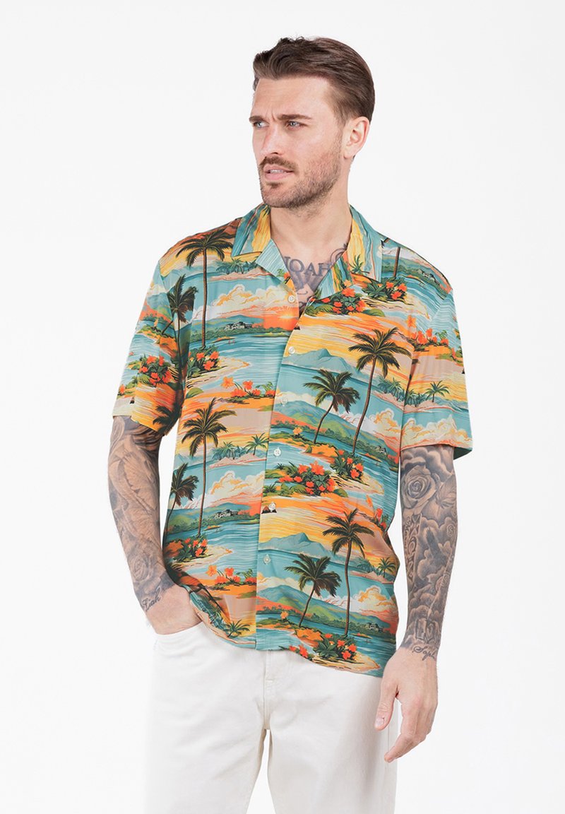 Homme tatoué portant une chemise à manches courtes aux imprimés tropicaux colorés et un pantalon blanc, regardant sur le côté avec les mains partiellement dans les poches.
