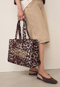 Geantă tote cu imprimeu leopard, inscripție aurie "VENICE", material textil, două mânere și formă structurată. Purtați cu pantaloni scurți bej.