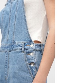 Denim-dress i lys blå, med glidelås foran, knapper på siden, og en teksturert hvit topp under. Enkel design med funksjonelle detaljer.