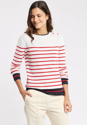 Femme aux cheveux foncés portant un pull blanc à rayures rouges avec des boutons sur l'épaule, un pantalon crème et un sous-pull noir.