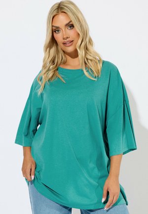 Lyshåret kvinde iført løs teal kortærmet skjorte og lyseblå jeans, stående mod en ensfarvet hvid baggrund.