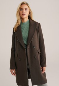 Cappotto di lana marrone a doppio petto con rever a punta, tasche frontali e bottoni neri, indossato sopra un maglione verde a trama.