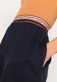 Detailní záběr osoby v námořnických kalhotách se značkovým elastickým pasem a oranžovém žebrovaném topu, ruka zasunutá v kapse.