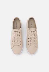 Paire de baskets beiges à lacets avec œillets dorés et semelles blanches, avec le logo Tommy Hilfiger sur la languette et le côté.