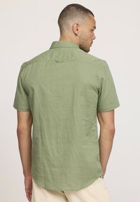 Chemise à manches courtes de couleur vert clair, fabriquée en tissu léger. Elle présente un col standard, une fente au dos et un ourlet légèrement arrondi.