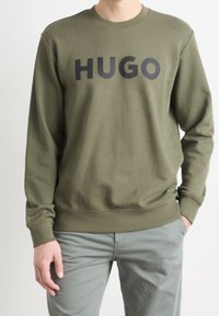 Olivově zelený svetr vyrobený z bavlny, s velkým černým logem "HUGO" na přední straně, s pružnými manžetami a lemem.