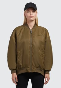 Olívovo zelená bomber bunda so zipsom, rebrovaným golierom a manžetami, so bočnými vreckami a hladkým, jemným materiálom. Nosená čierna čiapka.
