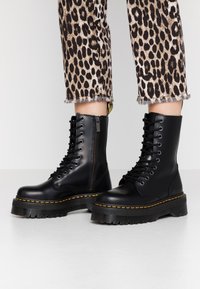 Botas negras de combate con cordones, puntera redonda, suela de plataforma de goma, costuras amarillas y detalle de cremallera lateral. Combinadas con jeans de estampado de leopardo.
