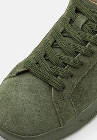 Gröna mockaskor med texturerad yta, försedd med gröna snören och en vadderad krage. Sulan är i gummi med lätt mönstring för grepp.