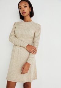 Robe en tricot beige avec texture côtelée, manches longues, encolure arrondie et ourlet légèrement évasé. Tissu doux et léger.