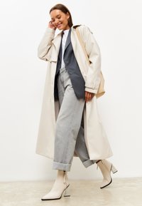 Beige trench coat päälle harmaa bleiseri, yhdistettynä vaaleanharmaisiin leveälahkeisiin farkkuihin ja kermavalkoisiin nilkkureihin, joissa on teksturoitu hopeanvärinen korko.