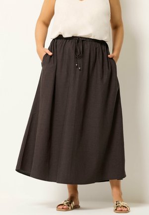 Kvinde iført en hvid ærmeløs top og en lang mørk nederdel med lommer, kombineret med gyldne slip-on sandaler, stående mod en ensfarvet baggrund.