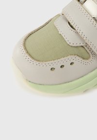 Gros plan sur l'avant d'une chaussure de sport en cuir beige, tissu vert clair à motif quadrillé, semelle verte avec des trous de ventilation et une fermeture à bande auto-agrippante.
