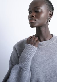 Jeune femme aux cheveux tressés portant un pull gris, tenant le col près de son cou, regardant sur le côté sur un fond uni.