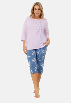 Kobieta stojąca boso, ubrana w jasnofioletową bluzkę z długim rękawem z motywem liści oraz niebieskie spodnie capri z pasującym wzorem liści.