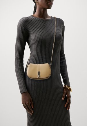 Sac en cuir beige à bandoulière avec une chaîne dorée, de forme courbée, et un accent en métal central. Porté sur une robe grise à côtes.