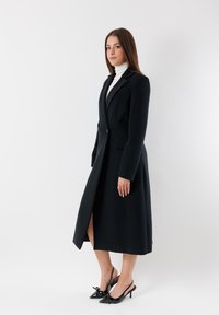 Cappotto lungo nero con silhouette su misura, caratterizzato da un design doppiopetto, spacchi laterali e abbinato a tacchi in pelle verniciata nera.