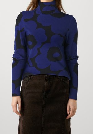 Femme portant un pull à col roulé bleu et noir à motifs floraux, associé à une jupe en denim marron foncé taille haute sur fond uni.