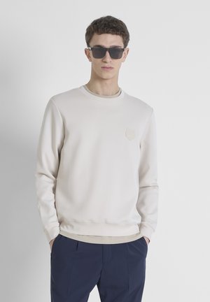 Sudadera beige con cuello redondo y un sutil parche de logo en el pecho, combinada con pantalones navy y gafas de sol oscuras.