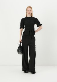 Svart bluse med pufferm og smocket tekstur, med peplum-kant, kombinert med høyhalsede svarte bukser og en liten svart veske.