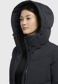 Puffjakke i mørkegrå med høj krave, stor hætte og lynlås. Har trykknapper og en quiltet struktur for isolation.