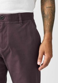 Burgunderfarbene Hose aus glattem, strukturiertem Stoff mit einem Knopfverschluss, seitlichen Taschen und einer taillierten Passform.